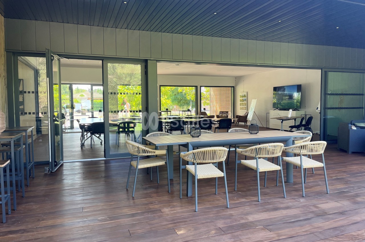 Salle moderne avec grandes baies vitrées, table en bois entourée de chaises et vue sur un espace lumineux extérieur.