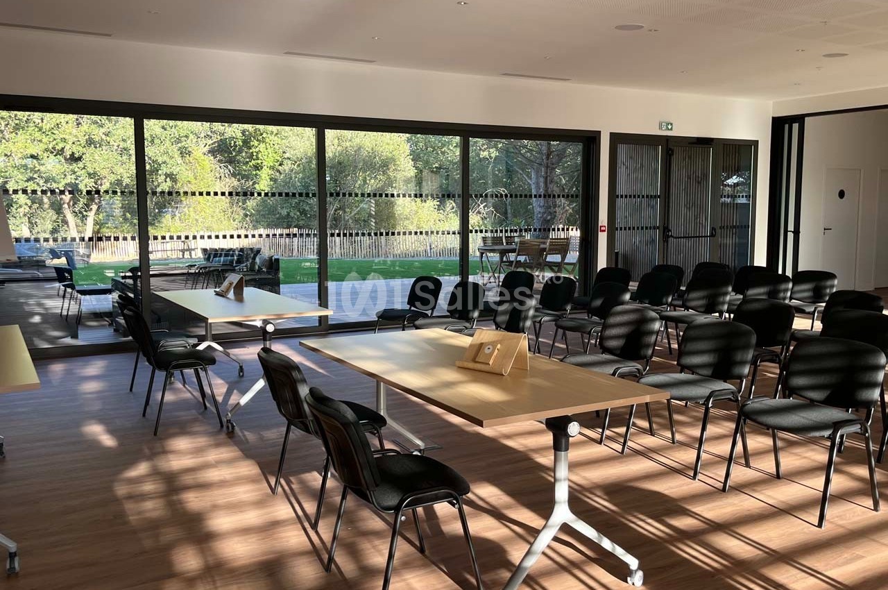 Salle lumineuse avec baies vitrées, aménagée avec des chaises et des tables, donnant sur un espace extérieur verdoyant.