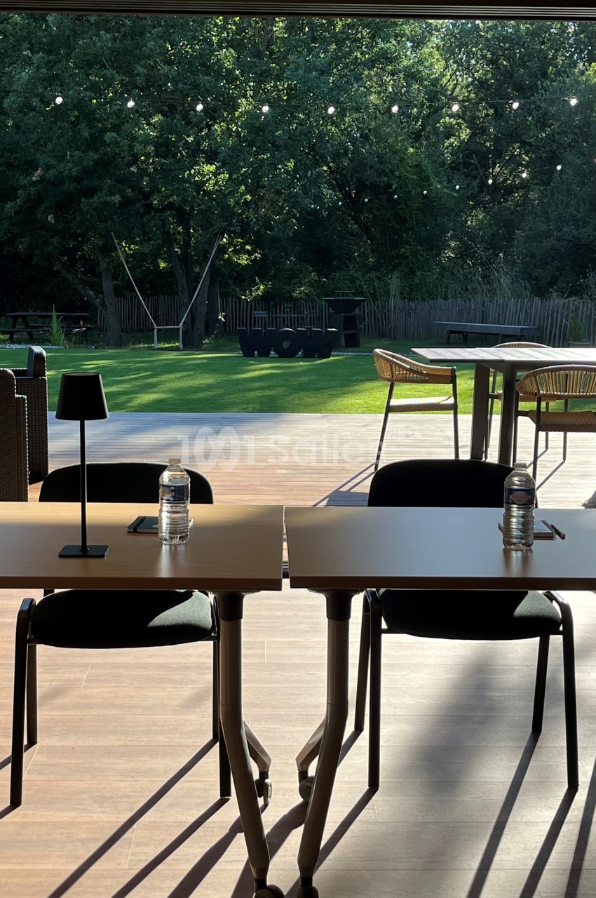 Deux tables avec chaises, lampes et bouteilles d'eau, devant une terrasse ensoleillée donnant sur un jardin verdoyant.
