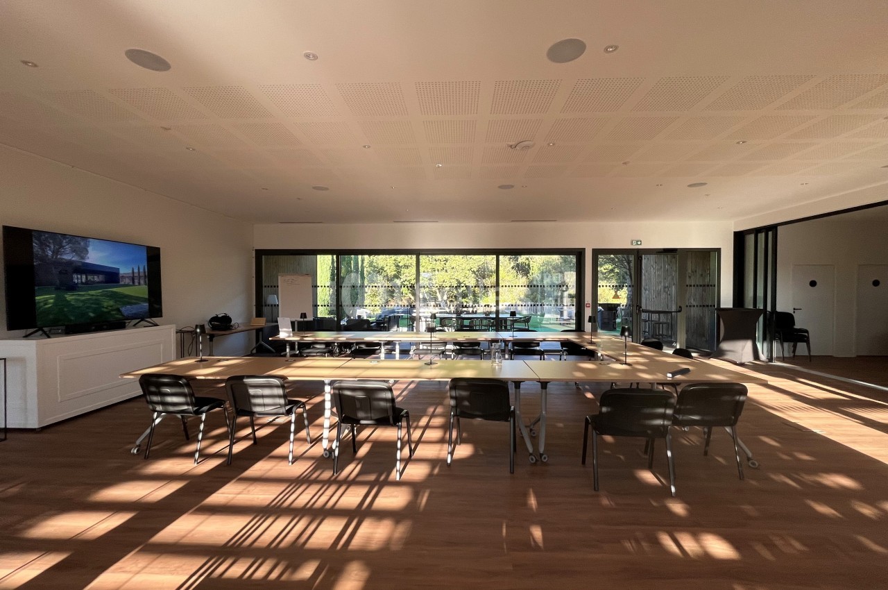 Salle de réunion lumineuse avec une grande table en bois, des chaises noires et une vue sur un jardin à travers des baies…