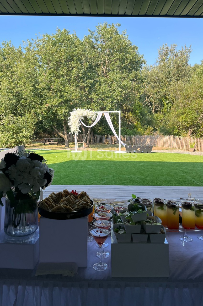Table de réception avec boissons, desserts et fleurs en premier plan, donnant sur un jardin avec arche décorative.