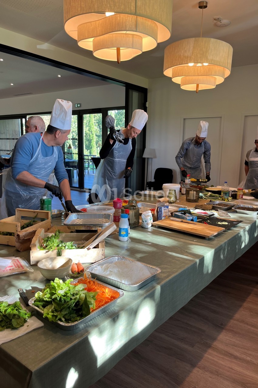 Des personnes en tabliers et toques cuisinent ensemble autour d'une grande table garnie d'ingrédients et d'ustensiles.