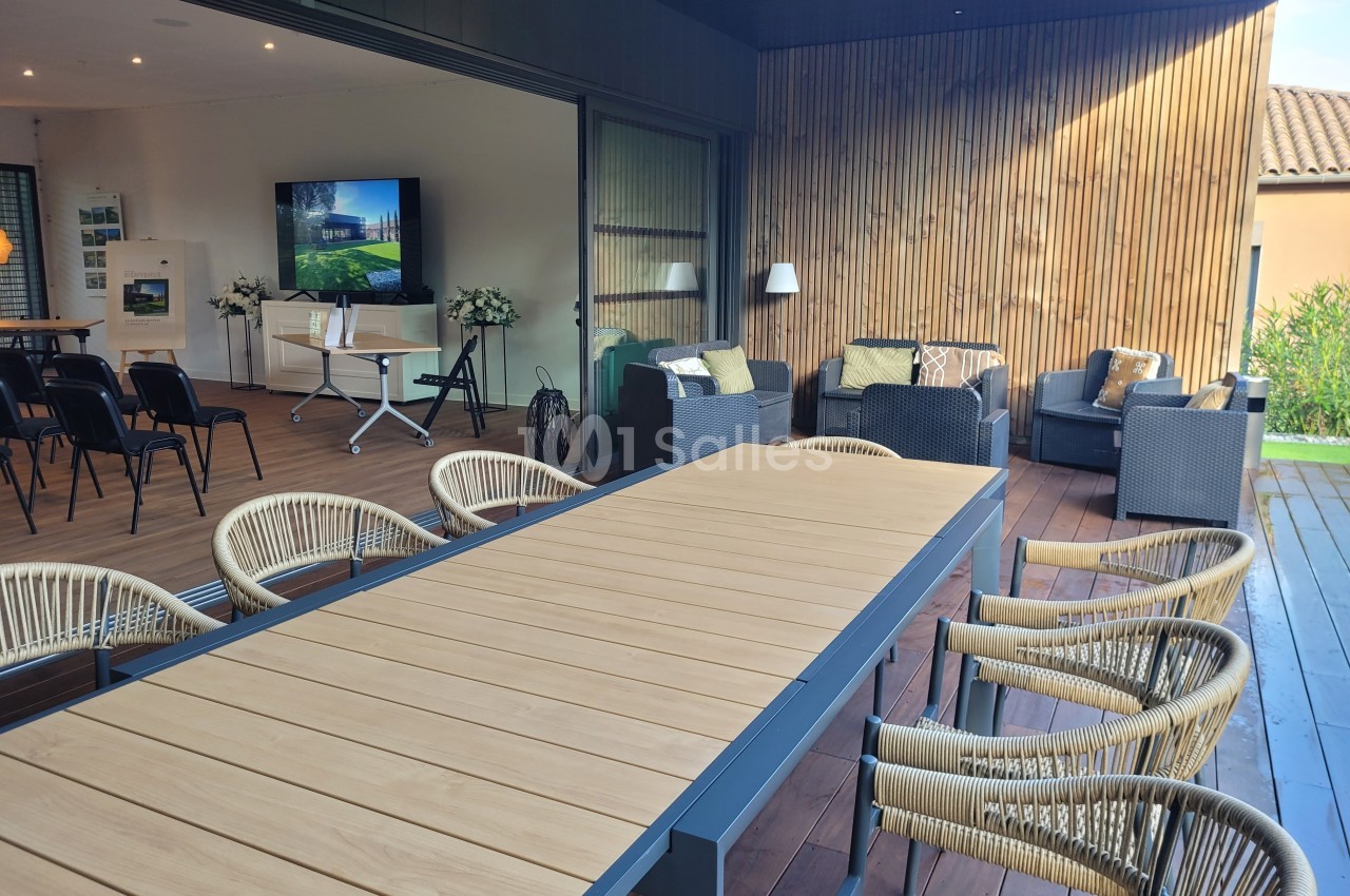 Terrasse aménagée avec une grande table en bois, des chaises en rotin et un coin salon avec fauteuils et écran en arrière…