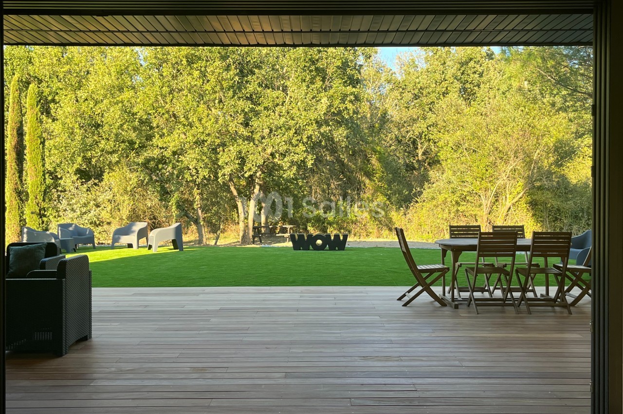 Vue sur un jardin avec pelouse, mobilier d'extérieur en bois et arbres en arrière-plan, depuis une terrasse en bois.