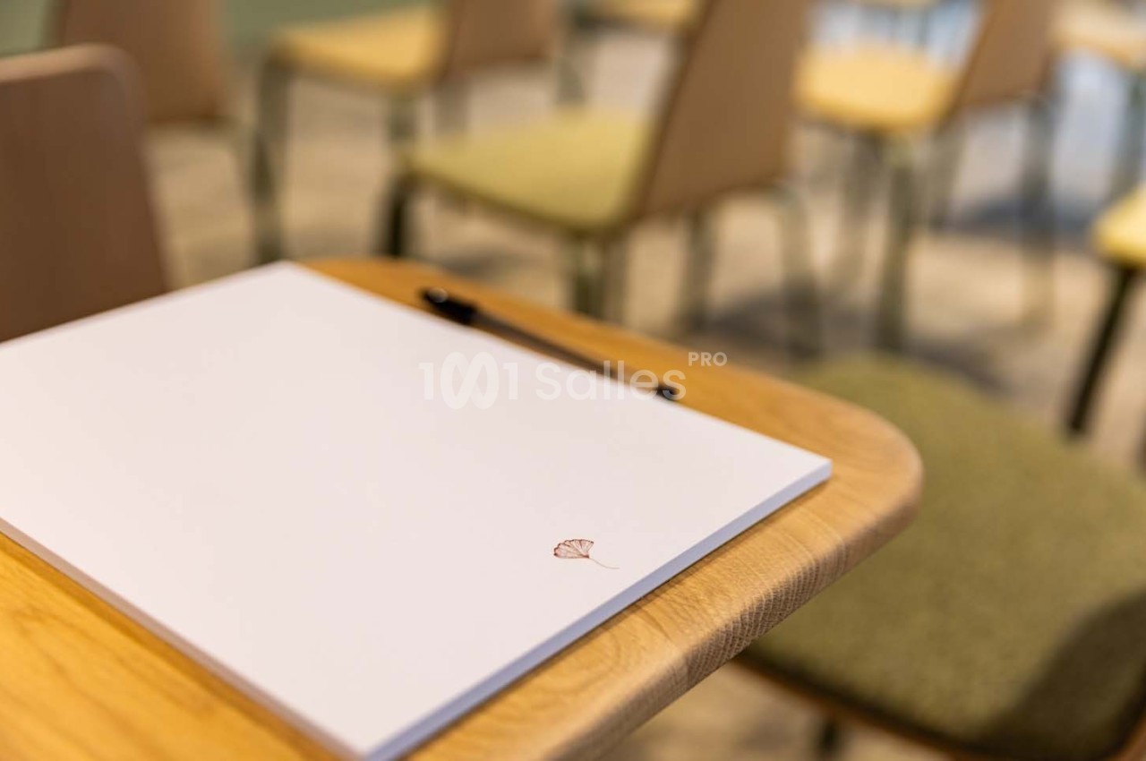 Bloc-notes blanc avec un stylo noir posé sur une table en bois, entouré de chaises floues en arrière-plan.