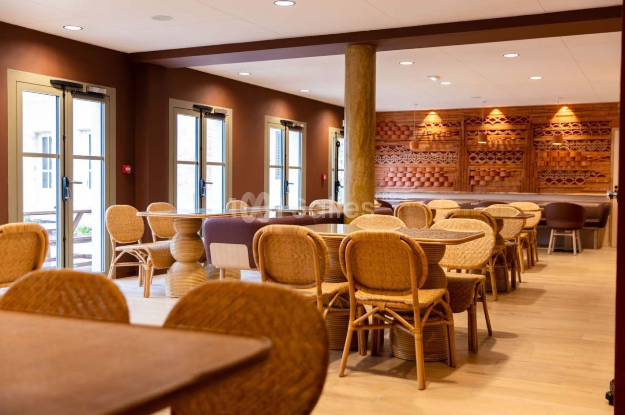 Salle lumineuse avec tables et chaises en rotin, décorée de boiseries murales et baignée de lumière naturelle.