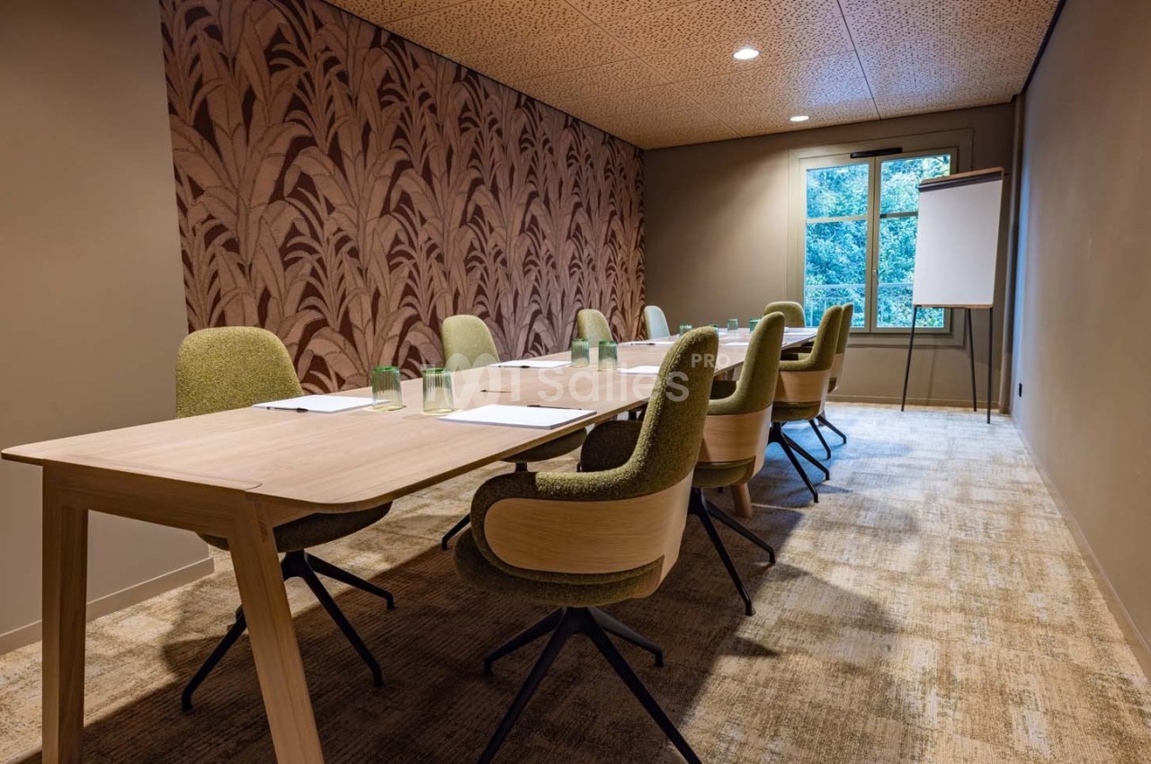 Salle de réunion avec table en bois, chaises vertes, tableau blanc et vue sur un extérieur arboré.