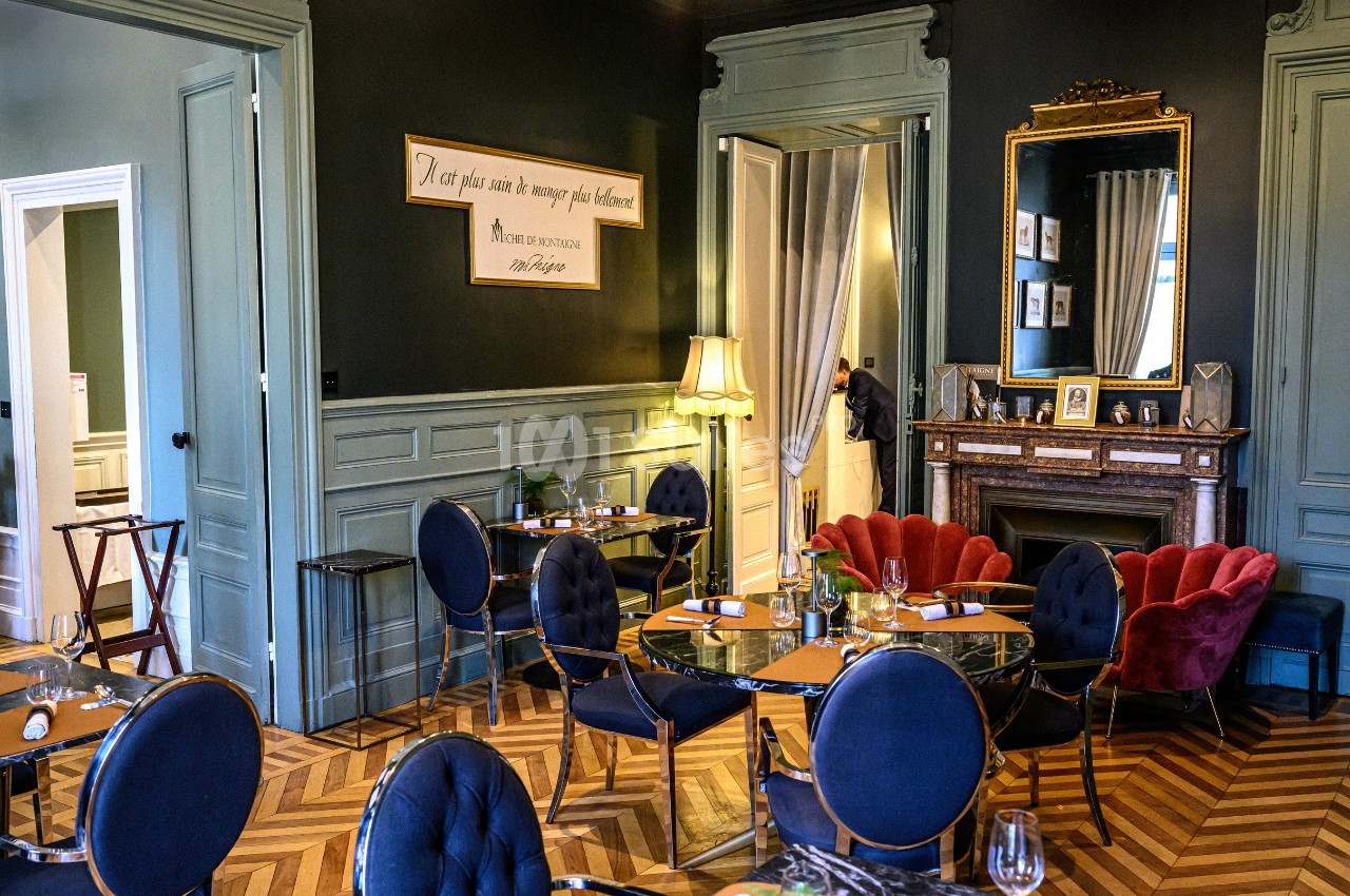 Salle de restaurant élégante avec parquet en chevrons, mobilier raffiné et cheminée ornée d'un miroir doré.