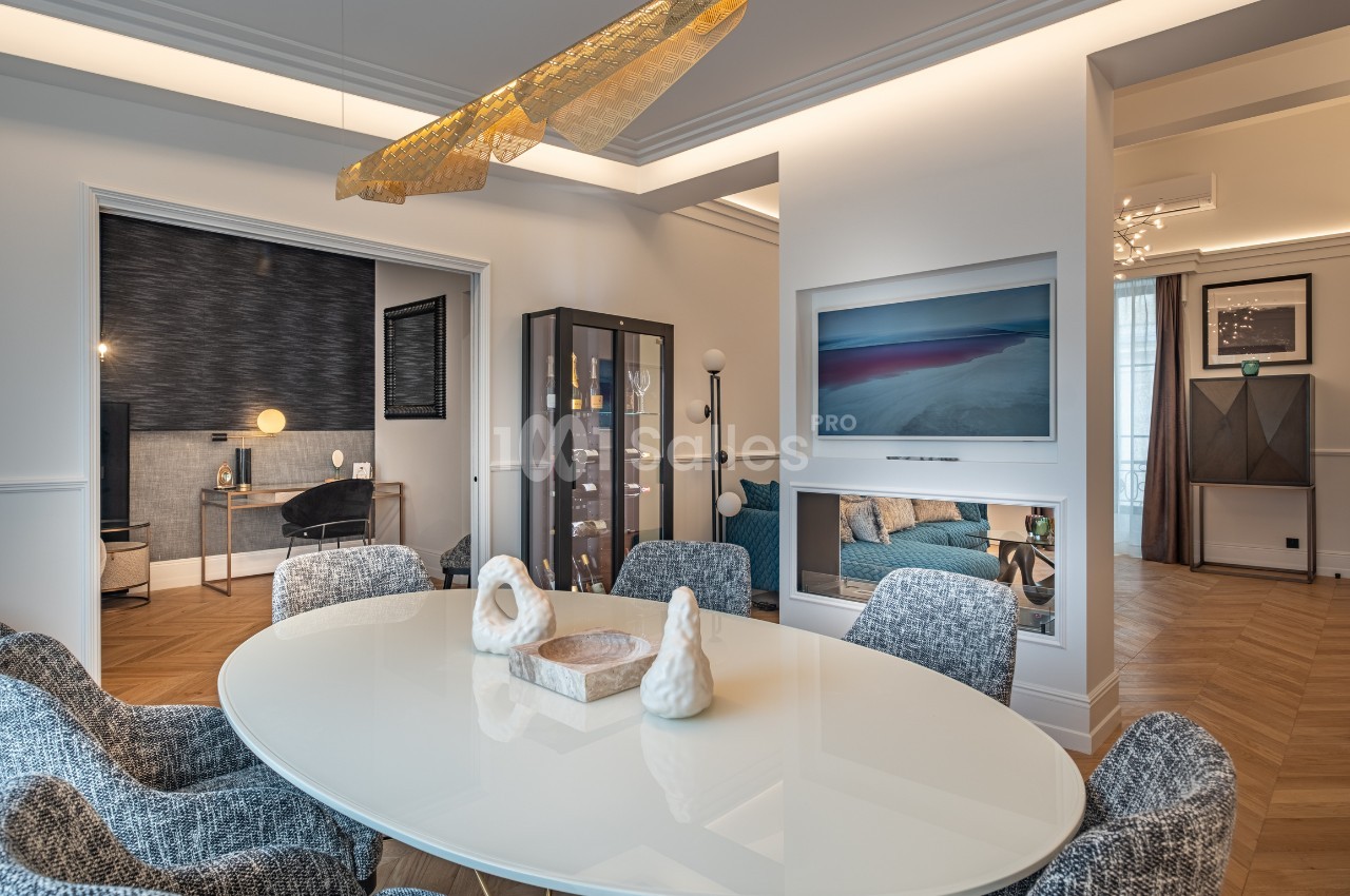 Salle à manger moderne avec table ovale blanche, chaises grises, éclairage design et vue sur un salon adjacent.
