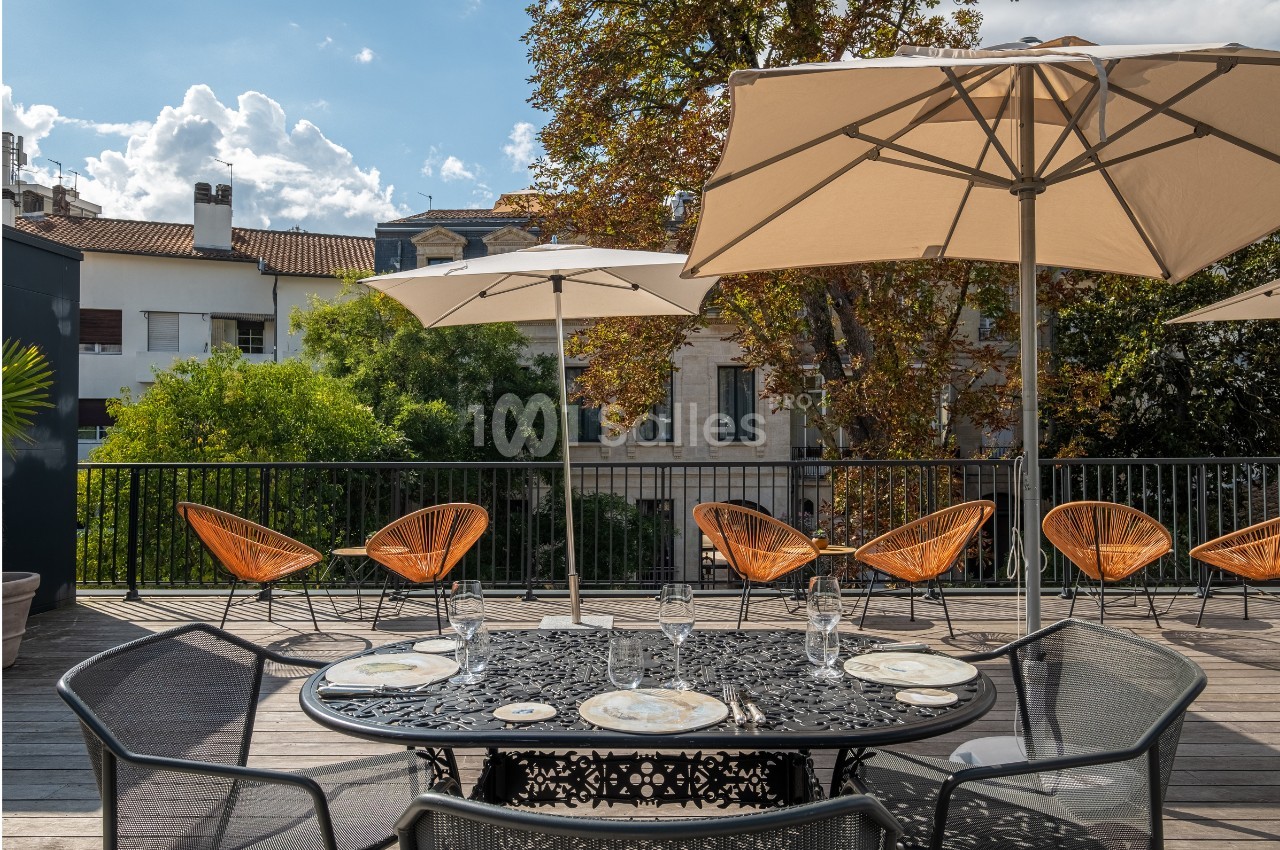 Terrasse ensoleillée avec table en fer forgé, chaises en rotin et parasols, entourée de verdure et bâtiments en arrière-plan.