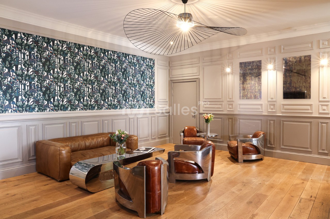 Salon élégant avec fauteuils en cuir, tables basses métalliques, parquet en bois clair et mur décoré de papier peint floral.