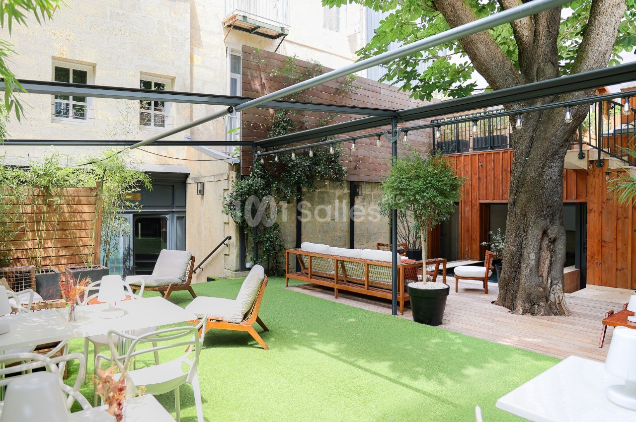 Cour extérieure aménagée avec mobilier de jardin, terrasse en bois, pelouse artificielle et grand arbre central.