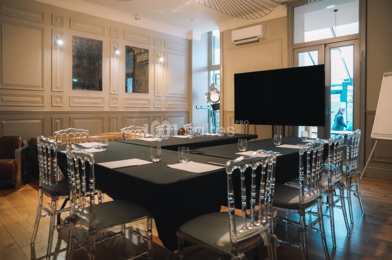 Salle de réunion élégante avec table en U, chaises transparentes, écran noir et éclairage tamisé.