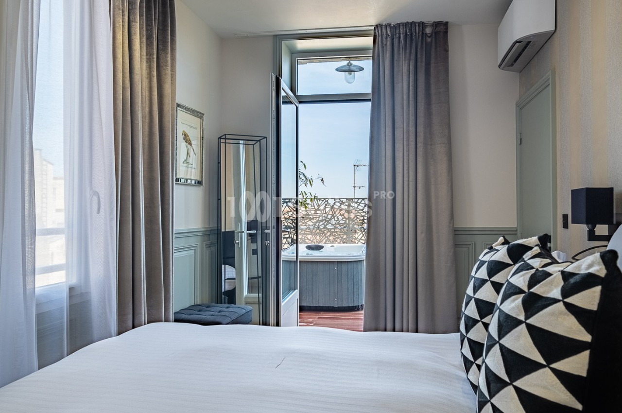 Chambre lumineuse avec lit double, rideaux gris, miroir, balcon ouvert et vue sur un jacuzzi extérieur.