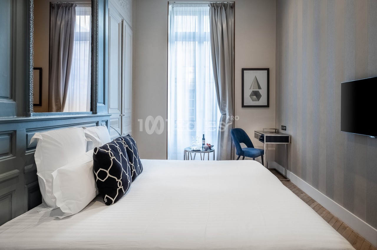 Chambre lumineuse avec un lit double, des coussins décoratifs, un bureau, une chaise et une grande fenêtre avec rideaux.