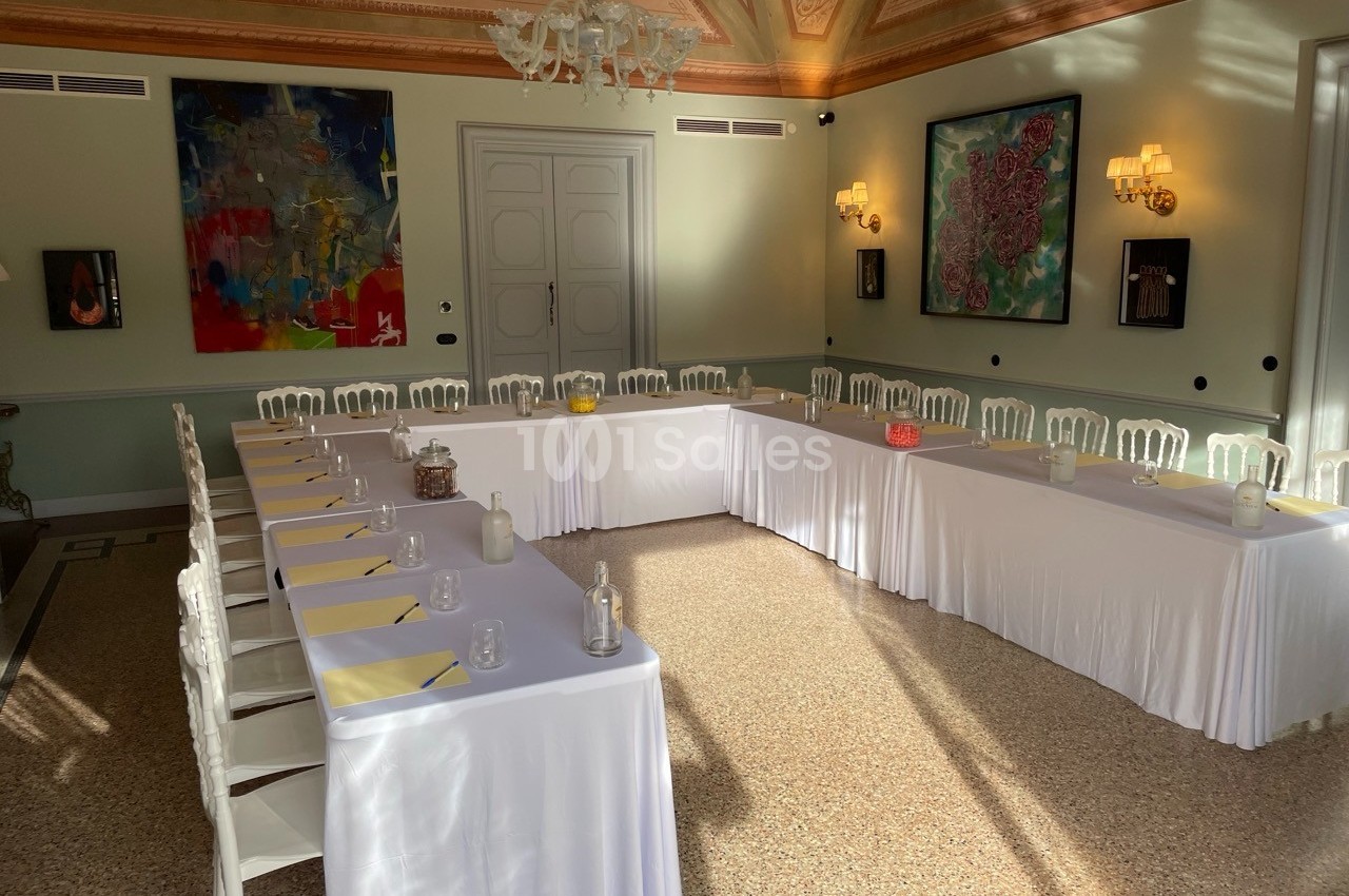 Salle de réunion lumineuse avec tables en U couvertes de nappes blanches, chaises blanches et œuvres d'art aux murs.