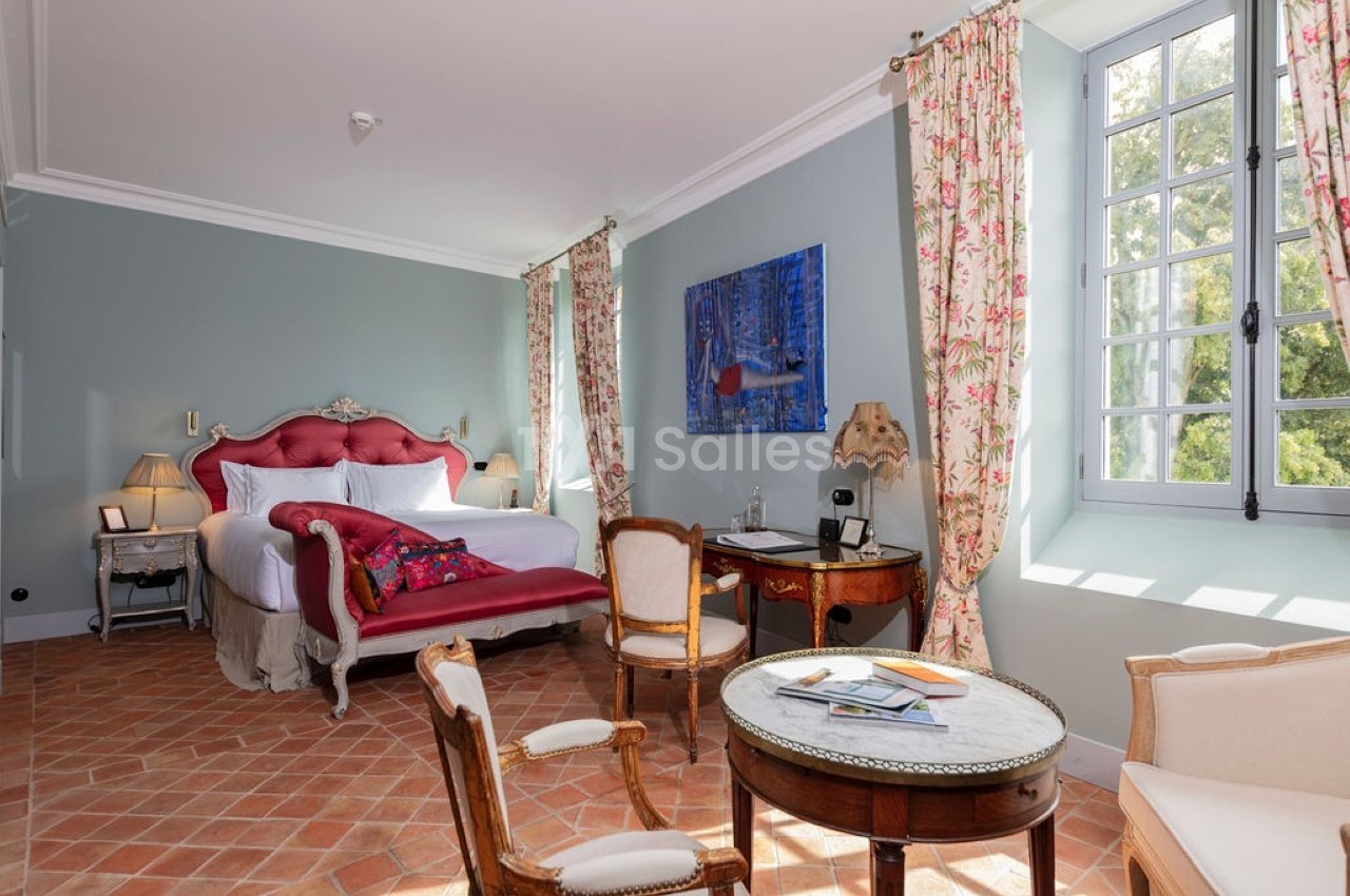 Chambre lumineuse avec lit double, fauteuils, table ronde, bureau, sol en terre cuite et fenêtre donnant sur un jardin.