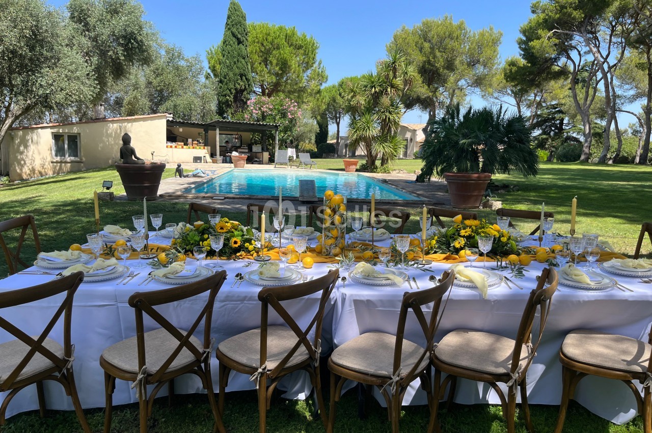 Table dressée en extérieur avec une décoration florale jaune, face à une piscine entourée de verdure et d'arbres.