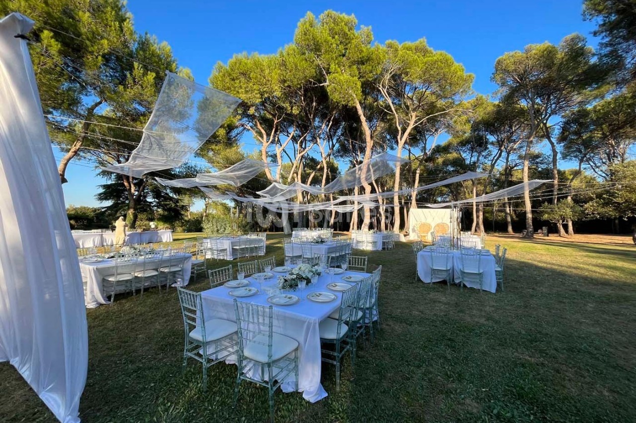 Tables dressées pour un événement en plein air sous des voilages blancs, entourées d'arbres et de pelouse.