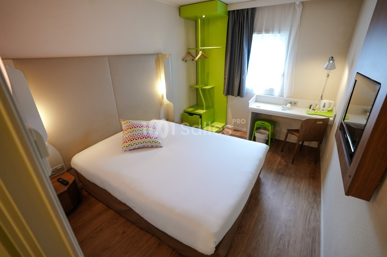 Chambre d'hôtel lumineuse avec lit double, bureau, chaise, étagères vertes et fenêtre avec rideaux.
