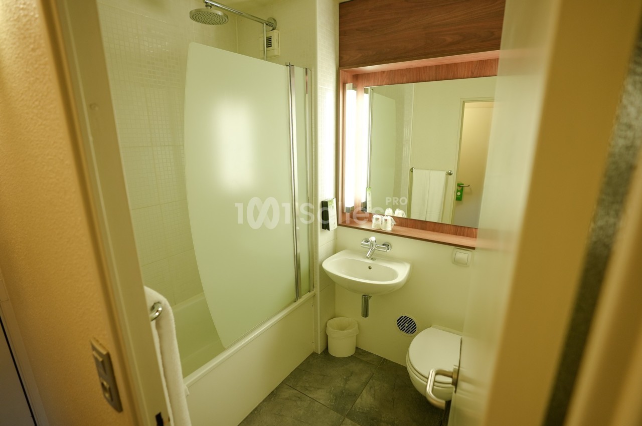 Salle de bain avec baignoire-douche, lavabo, miroir éclairé, toilettes et carrelage au sol.