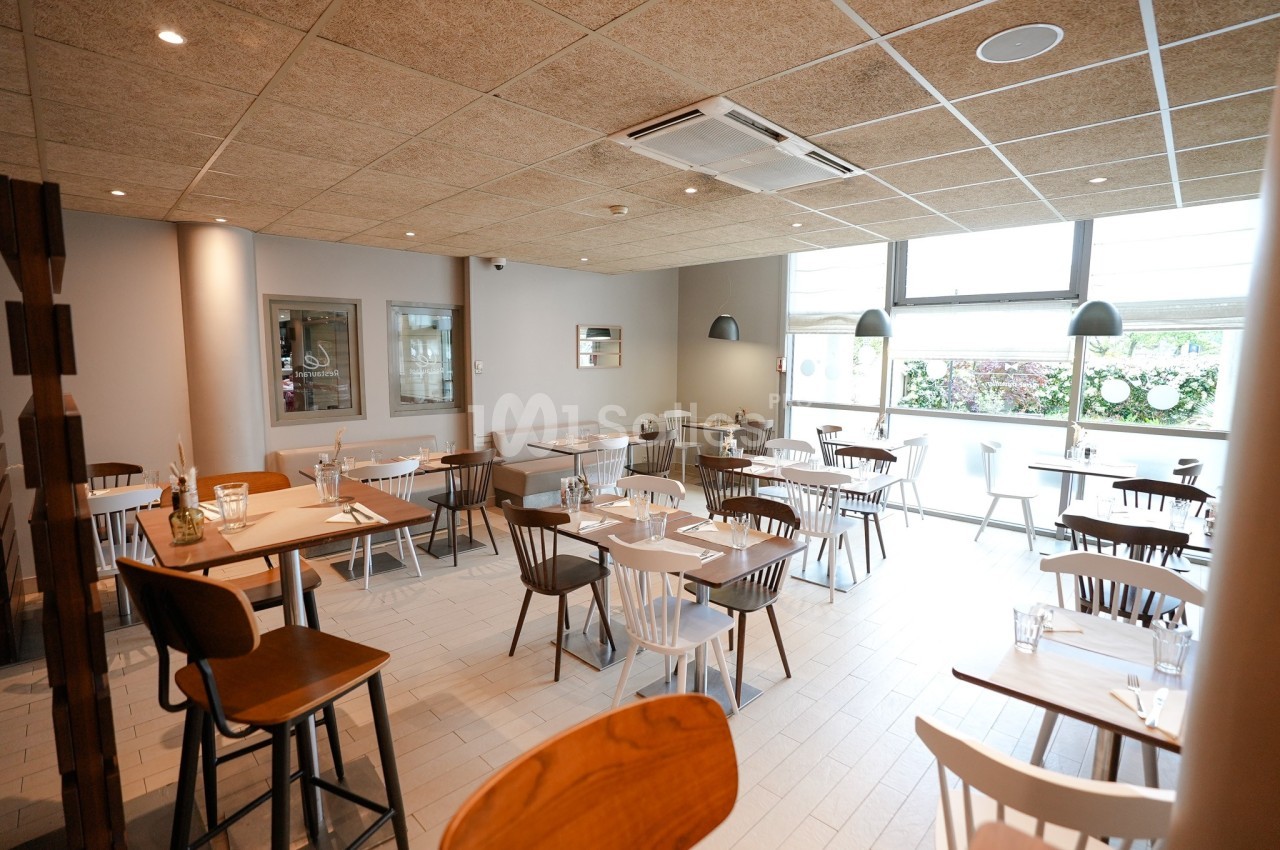 Salle de restaurant lumineuse avec des tables en bois clair, des chaises variées et une grande fenêtre donnant sur l…