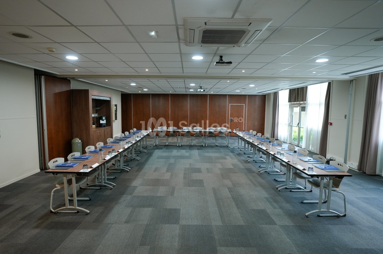 Salle de réunion spacieuse avec disposition en U, tables équipées de blocs-notes et chaises, éclairée par des fenêtres.