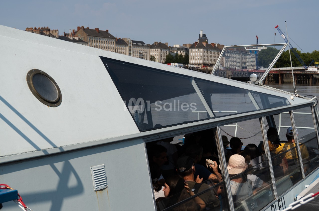 Bateau touristique avec passagers à bord, naviguant sur un fleuve près d'une ville avec des bâtiments en arrière-plan.
