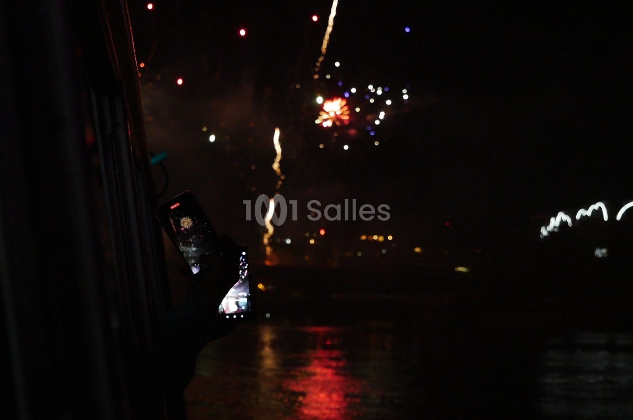 Une personne photographie un feu d'artifice nocturne avec un smartphone, près d'une surface d'eau réfléchissante.