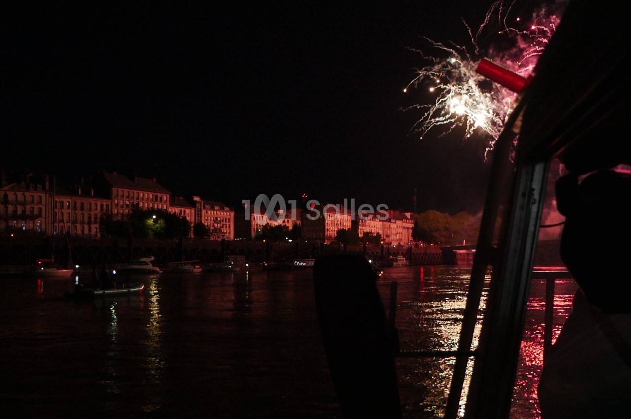 Feu d'artifice illuminant le ciel nocturne au-dessus d'une rivière bordée de bâtiments et de bateaux.