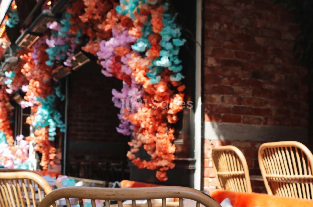 Façade en briques ornée de guirlandes de fleurs colorées avec des chaises en rotin au premier plan.