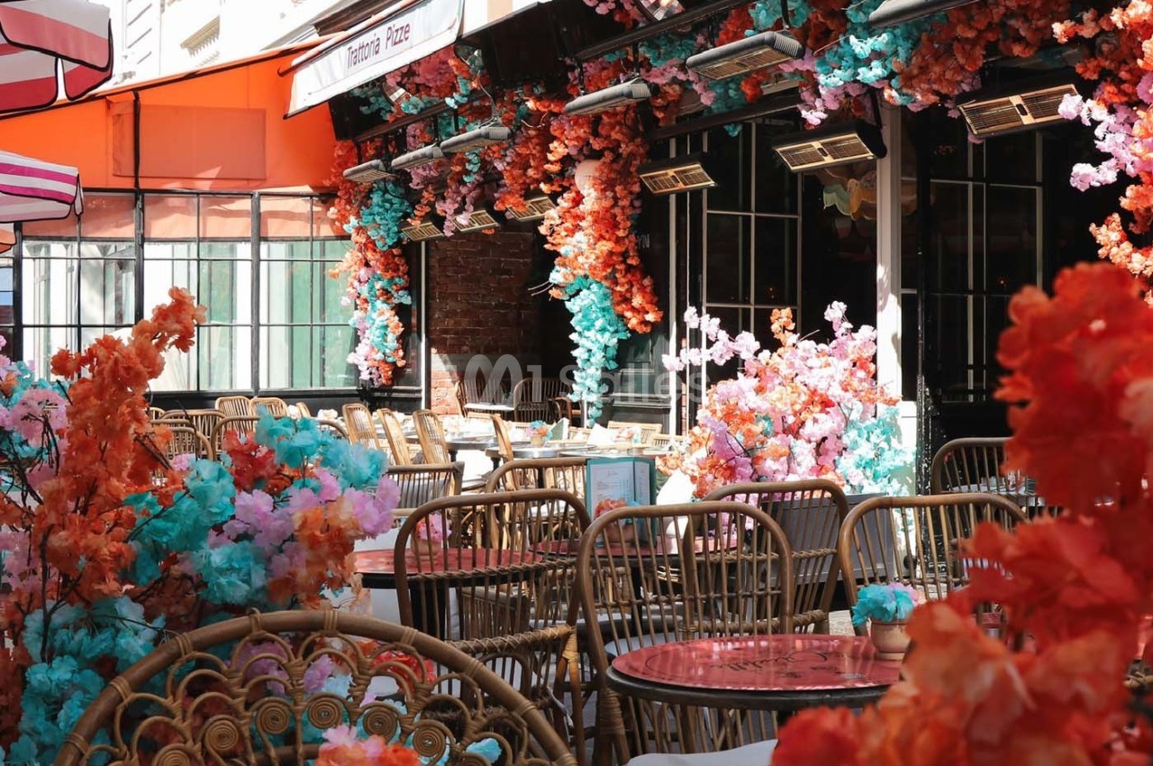 Terrasse de café décorée de fleurs colorées, avec des tables en rotin et une lumière naturelle abondante.