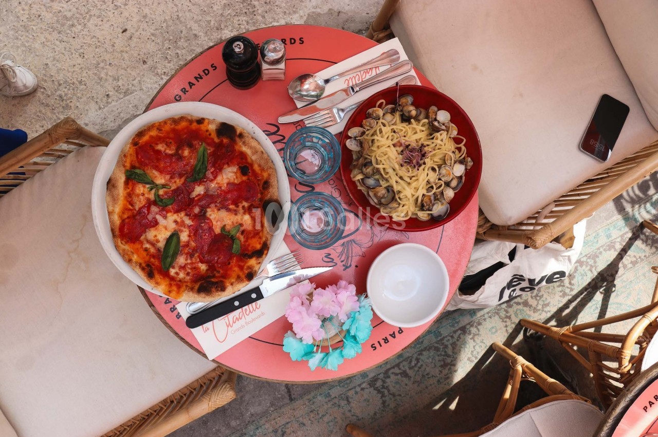 Table ronde avec une pizza garnie de basilic, un plat de pâtes aux palourdes, des verres et des couverts.