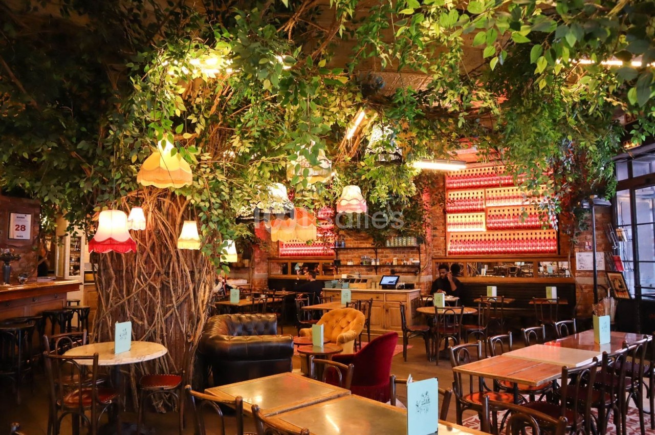 Salle de restaurant décorée avec des arbres, des lampes suspendues et un mobilier éclectique en bois et velours.