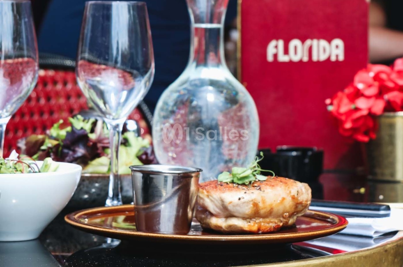 Plat de viande grillée avec garniture, posé sur une table de restaurant avec carafe d'eau, verres et décor rouge.