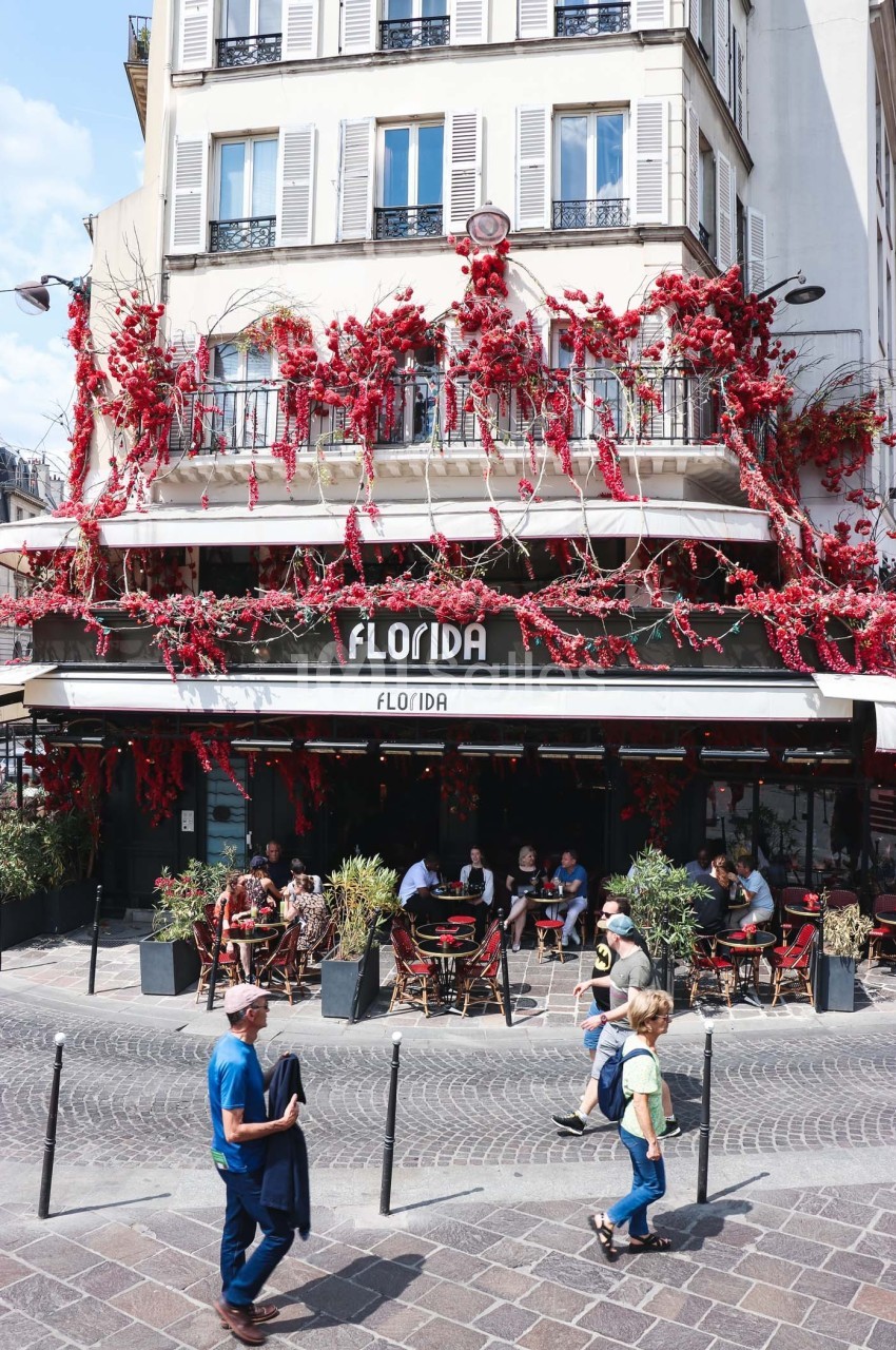 Façade d'un café avec terrasse, ornée de fleurs rouges, des passants marchent sur une rue pavée au premier plan.