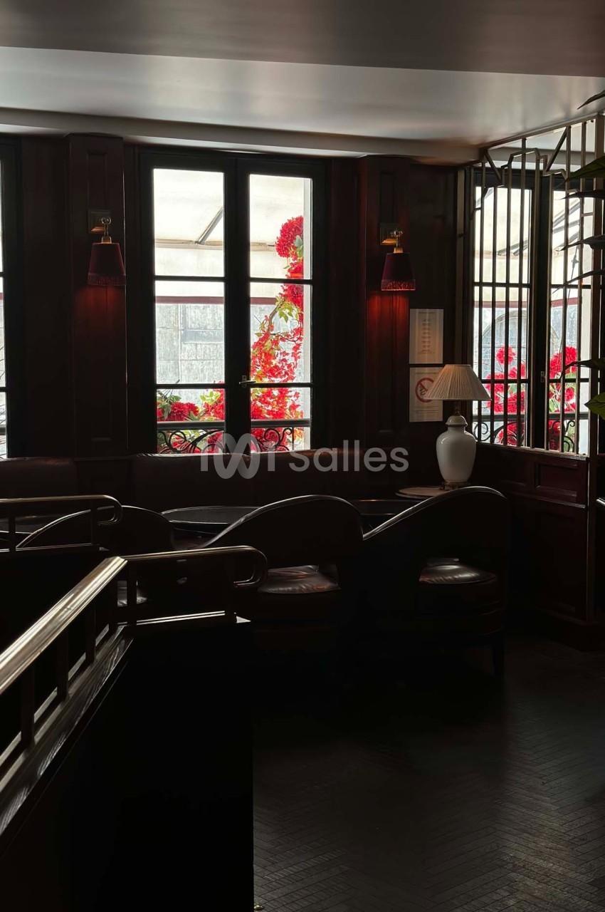 Intérieur sombre d'un café avec des banquettes, des fenêtres donnant sur des fleurs rouges et une lampe allumée.