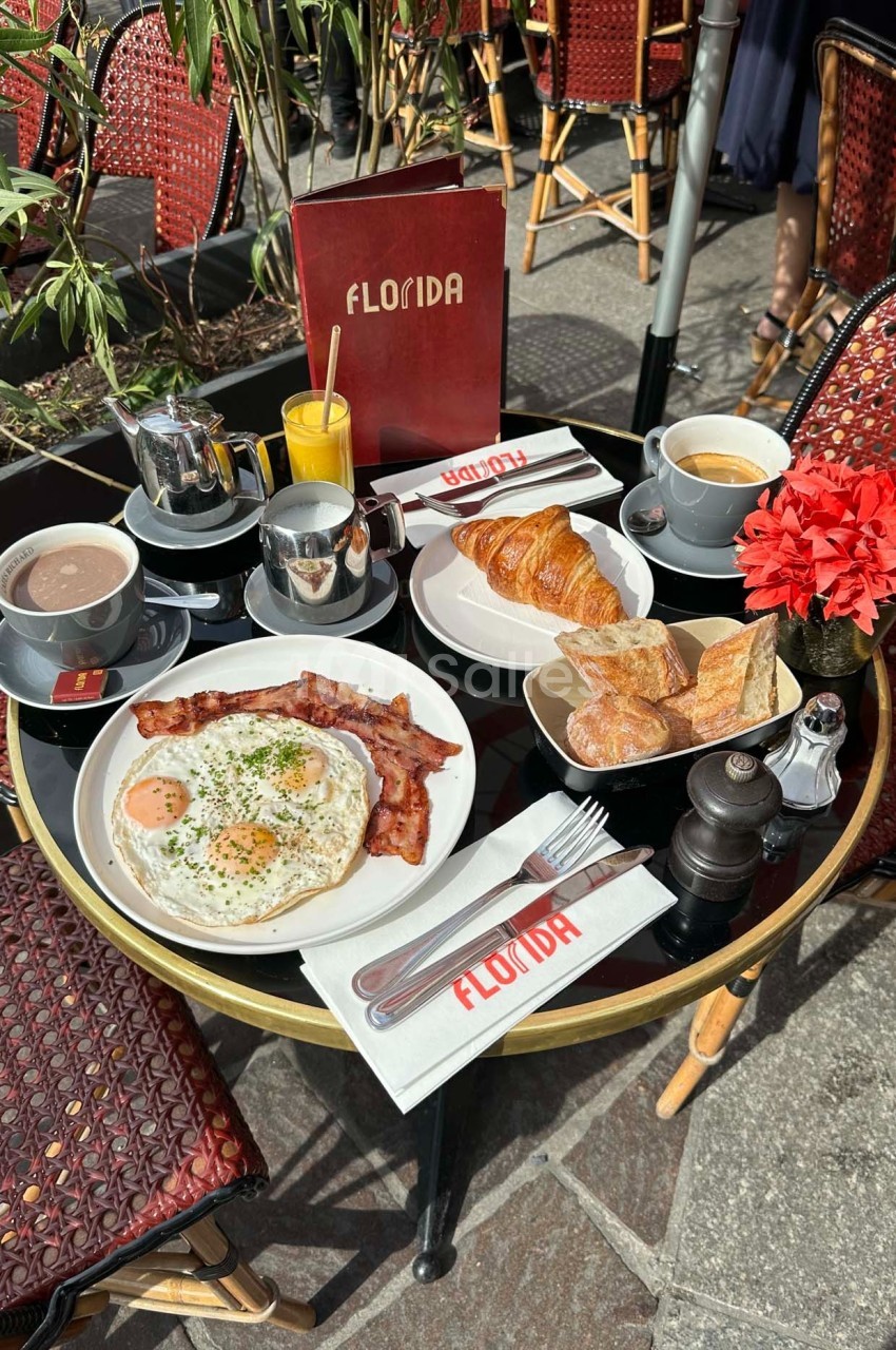 Petit-déjeuner en terrasse avec œufs, bacon, croissants, pain, boissons chaudes et jus d'orange sur une table ronde.