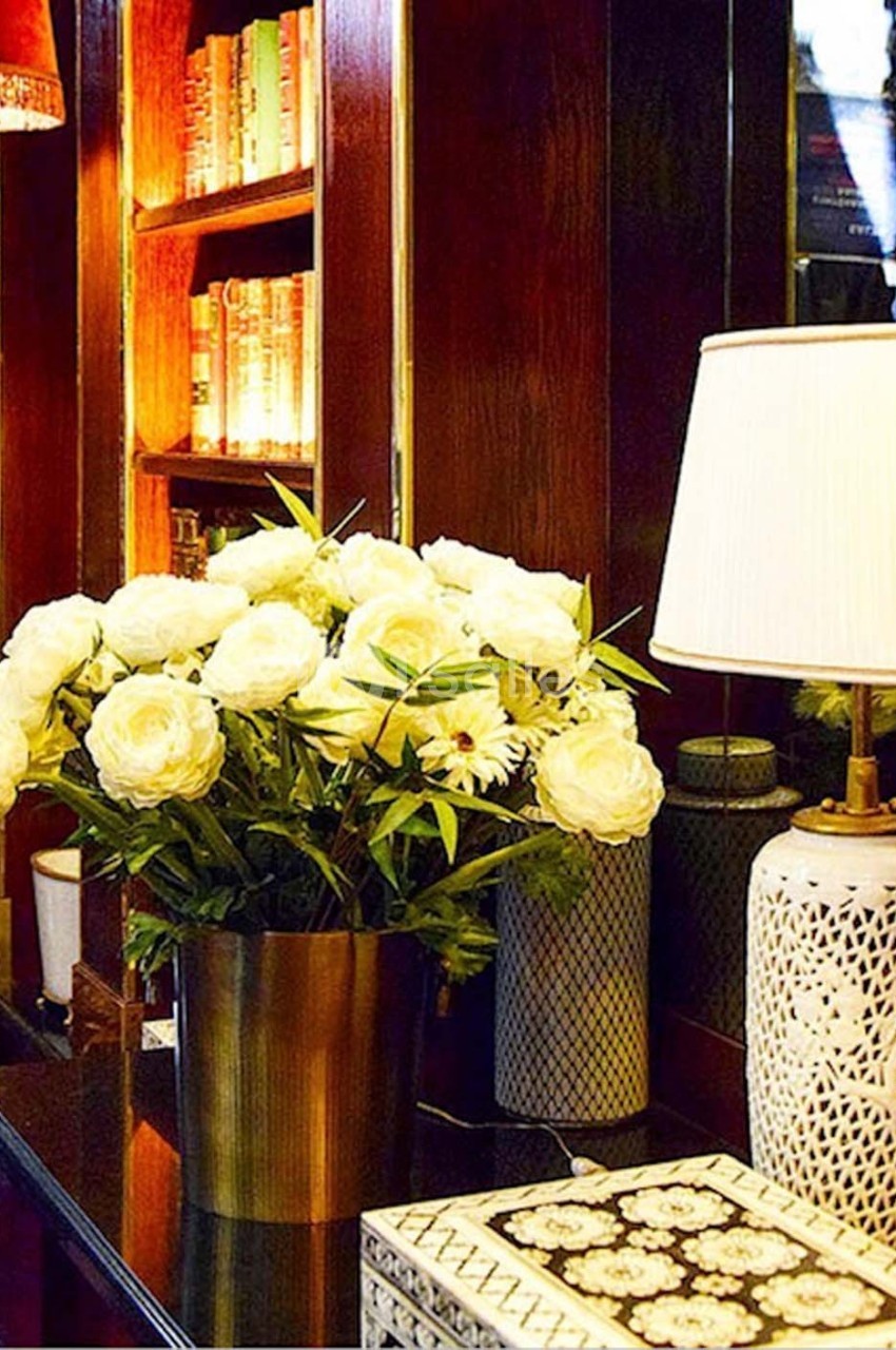 Bouquet de fleurs blanches dans un vase doré sur une table, avec une lampe et des étagères en arrière-plan.