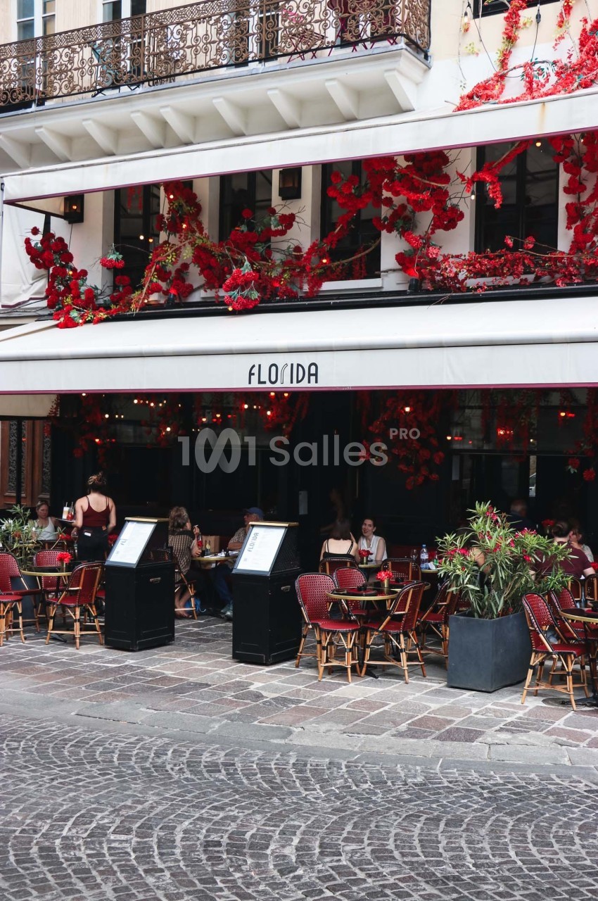 Terrasse d’un café-restaurant avec des tables occupées, façade ornée de fleurs rouges et enseigne ’Florida’.
