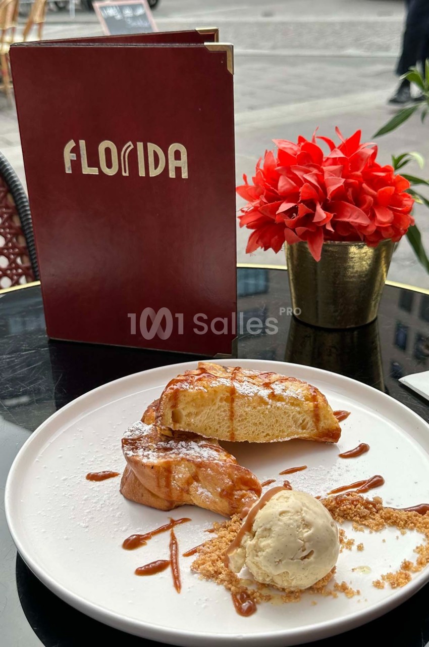 Assiette de pain perdu avec boule de glace et caramel, servie sur une table en terrasse avec un menu et une plante rouge.