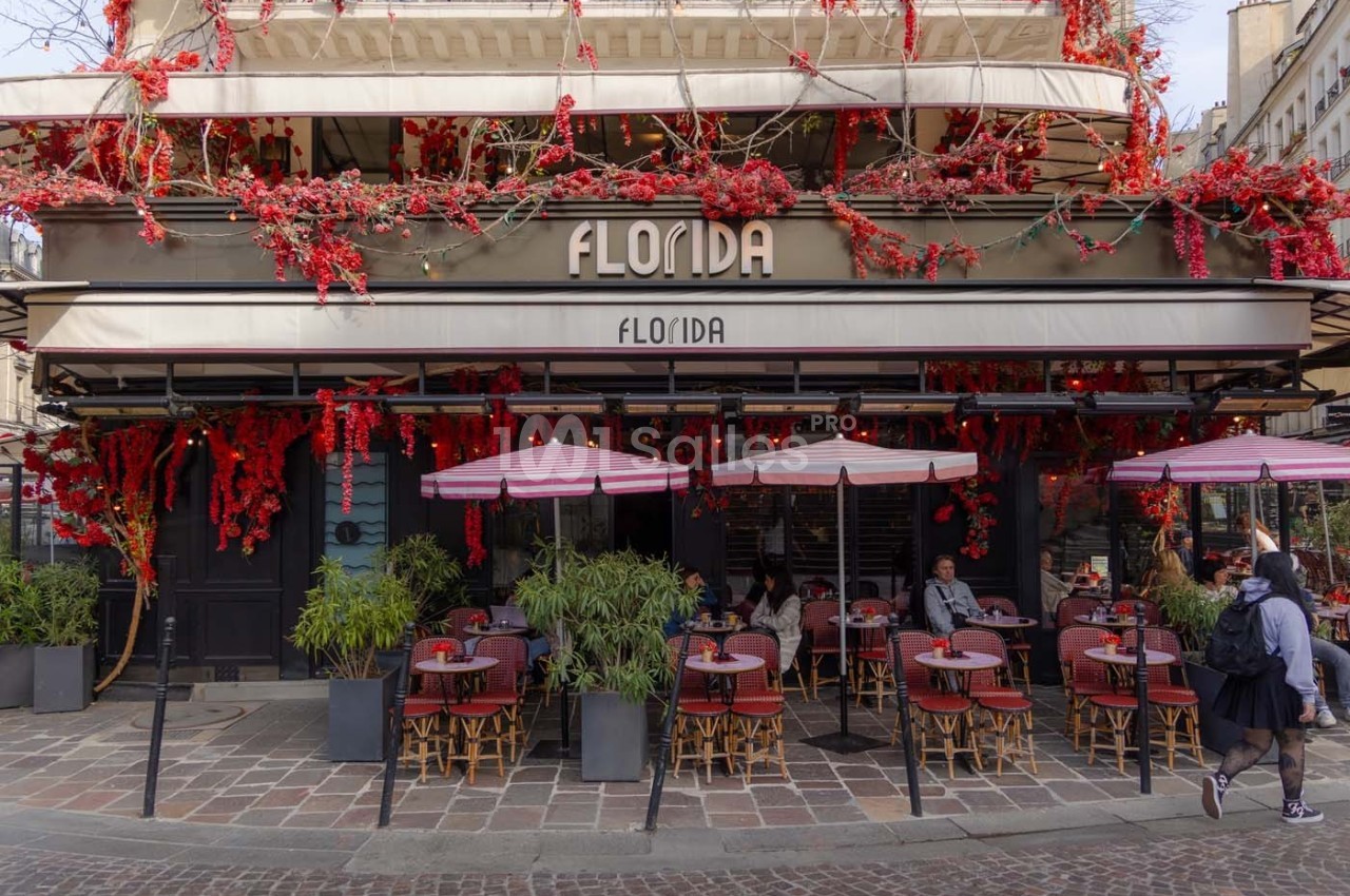 Façade d'un café avec terrasse, décorée de fleurs rouges, tables et chaises disposées sur un trottoir pavé.