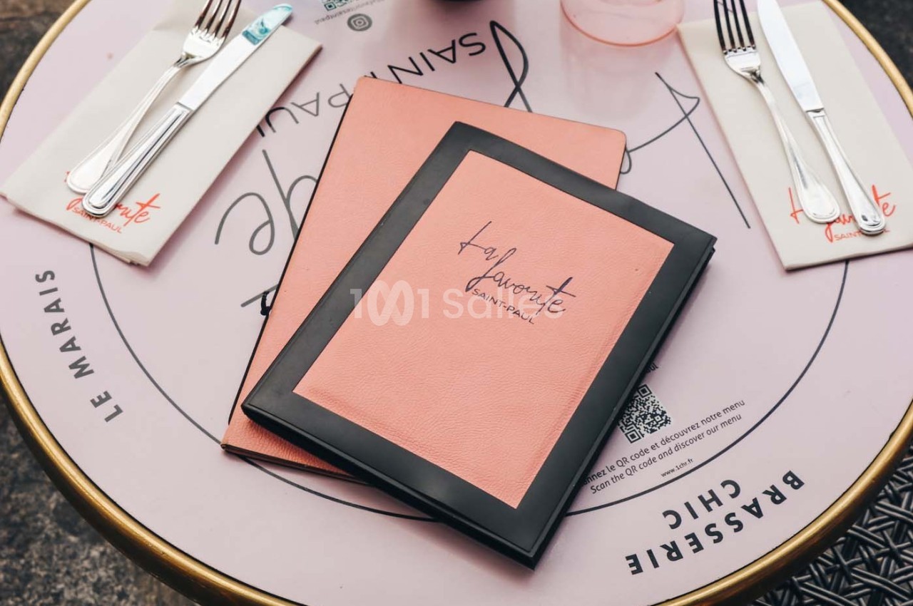 Deux menus roses posés sur une table ronde avec des couverts et des serviettes imprimées.