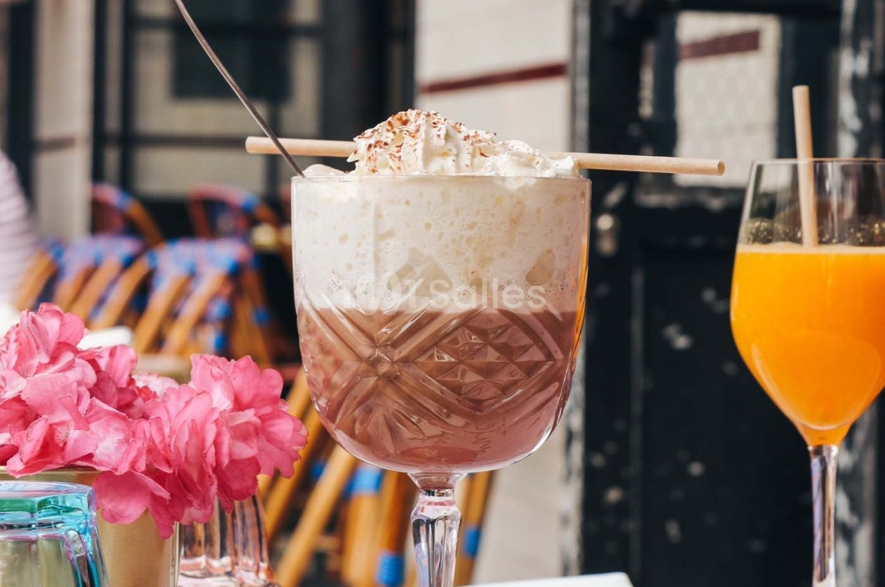 Verre de chocolat chaud avec chantilly et décoration, posé sur une table près de fleurs roses et un jus d'orange.