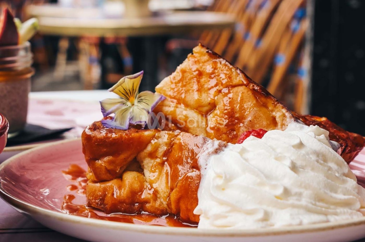 Tranche de pain perdu garnie de chantilly, fleur comestible et sirop, servie dans une assiette rose.