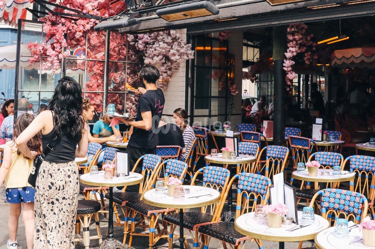Terrasse d'un café avec des tables colorées, des clients assis et des serveurs en activité sous des décorations florales.