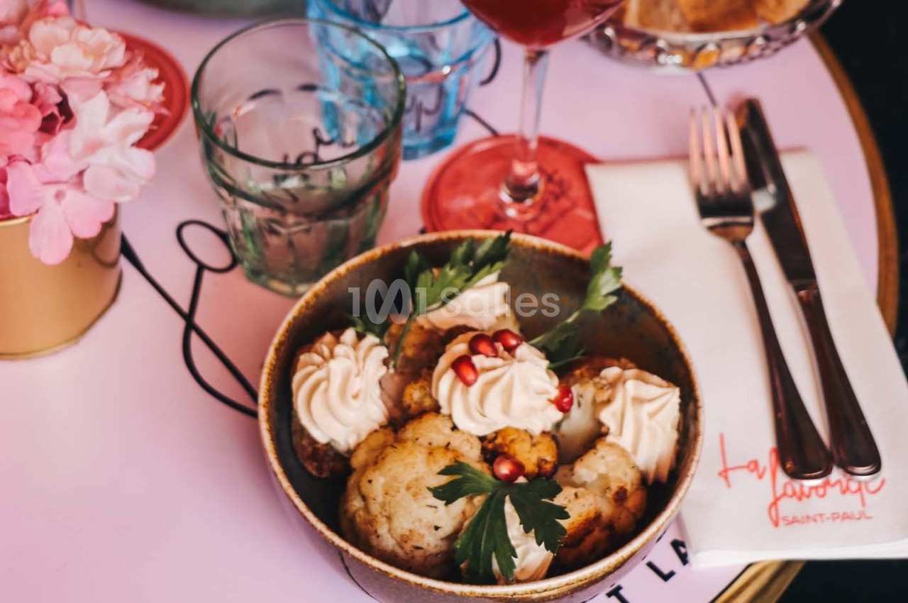 Bol de chou-fleur rôti garni de crème, graines de grenade et persil, posé sur une table avec couverts et verre.