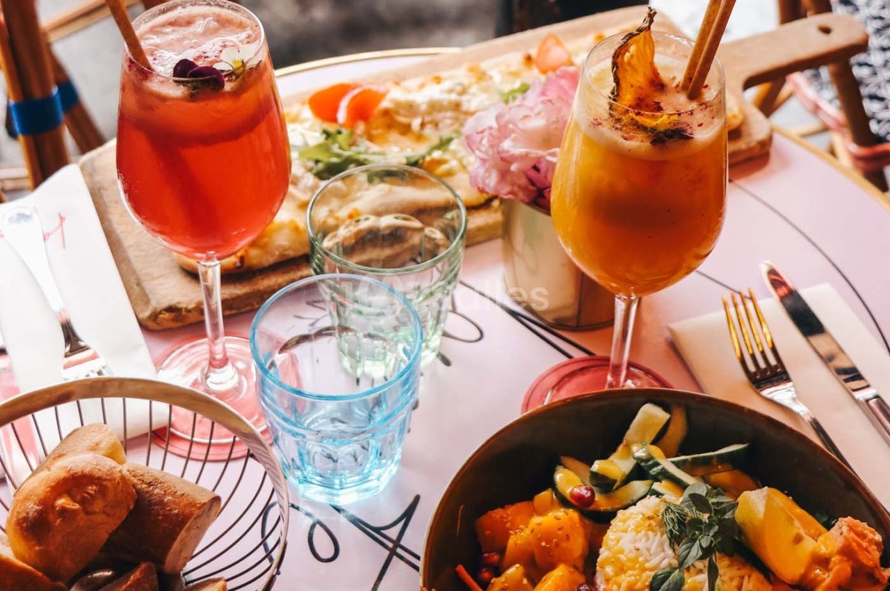 Deux cocktails colorés, une assiette de salade et une pizza sur une table dressée avec des verres et des couverts.