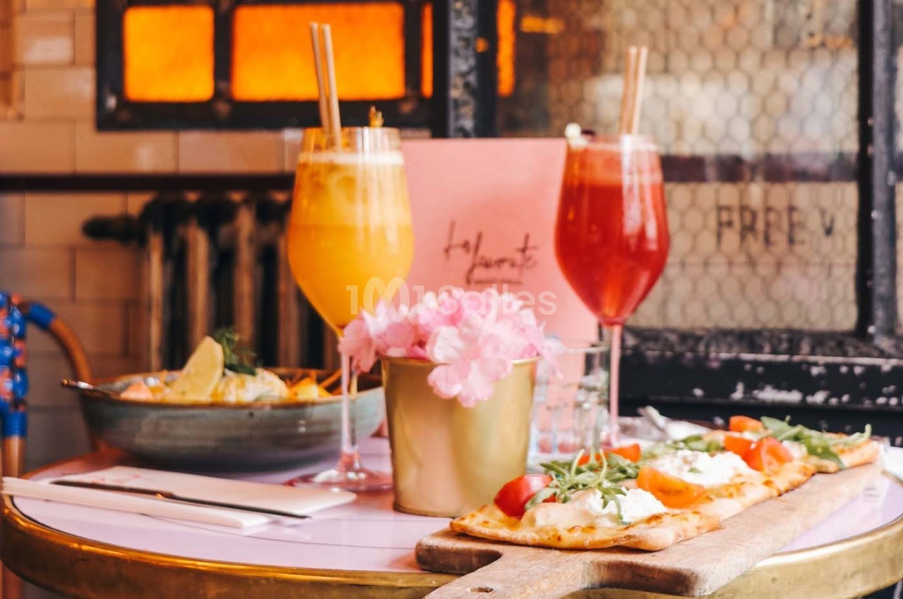 Boissons colorées, fleurs roses et plats de pizza sur une table dans un cadre chaleureux avec un four en arrière-plan.