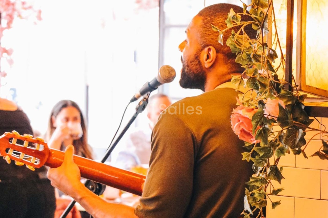 Un musicien joue de la guitare et chante au micro dans un café décoré de fleurs, devant un public attentif.
